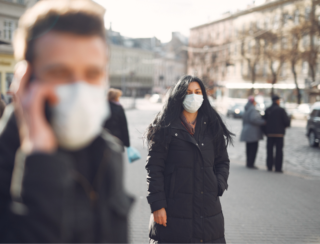 Man in medical mask in foreground on the phone blurred with focus on woman wearing a mask, both are outdoors in a city.
