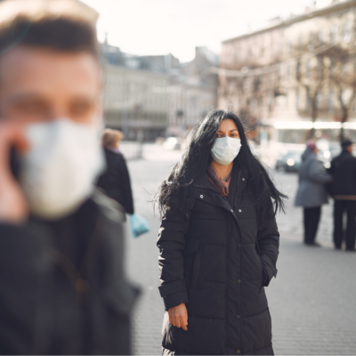 Man in medical mask in foreground on the phone blurred with focus on woman wearing a mask, both are outdoors in a city.